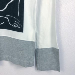 K&ouml;nnte beinhalten: Wei&szlig;es Langarmshirt mit grauem Saum und B&uuml;ndchen. Das Shirt hat eine schwarz-wei&szlig;e Grafik auf der oberen linken Seite, eine abstrakte Illustration einer Hand. Die &Auml;rmel sind wei&szlig;.