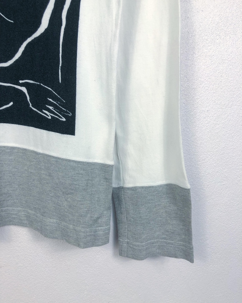 K&ouml;nnte beinhalten: Wei&szlig;es Langarmshirt mit grauem Saum und B&uuml;ndchen. Das Shirt hat eine schwarz-wei&szlig;e Grafik auf der oberen linken Seite, eine abstrakte Illustration einer Hand. Die &Auml;rmel sind wei&szlig;.