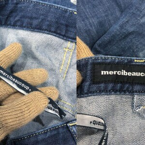 Op de afbeelding: Close-up van een blauwe denim jeans met gele stiksels. De jeans heeft een zwart label met de tekst "merci beaucoup," en een stoffen label met dezelfde tekst. Het stoffen label wordt vastgehouden door een gehandschoende hand.