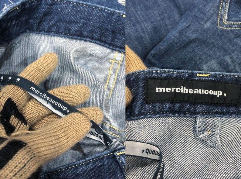 Op de afbeelding: Close-up van een blauwe denim jeans met gele stiksels. De jeans heeft een zwart label met de tekst "merci beaucoup," en een stoffen label met dezelfde tekst. Het stoffen label wordt vastgehouden door een gehandschoende hand.