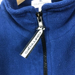 May include: A close-up of a navy blue fleece jacket with a black zipper. The jacket has a high collar and a tag with the brand name "courreges" in white text.