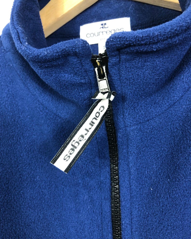 May include: A close-up of a navy blue fleece jacket with a black zipper. The jacket has a high collar and a tag with the brand name "courreges" in white text.