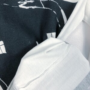 K&ouml;nnte beinhalten: Wei&szlig;es Langarmshirt mit einem schwarzen Grafikdesign. Das Design scheint eine abstrakte Landschaft oder Stadtlandschaft zu sein. Der &Auml;rmel des Shirts ist sichtbar und zeigt die Textur und die N&auml;hte des Stoffes.