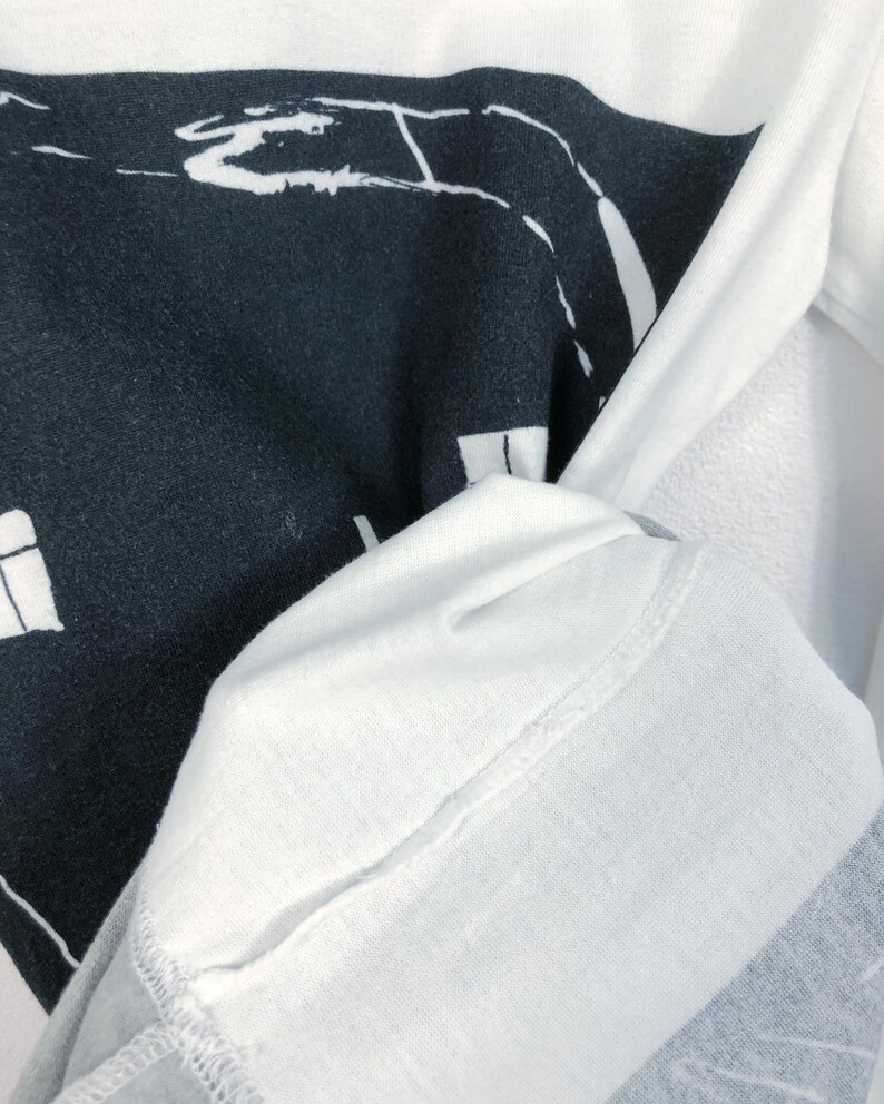 K&ouml;nnte beinhalten: Wei&szlig;es Langarmshirt mit einem schwarzen Grafikdesign. Das Design scheint eine abstrakte Landschaft oder Stadtlandschaft zu sein. Der &Auml;rmel des Shirts ist sichtbar und zeigt die Textur und die N&auml;hte des Stoffes.