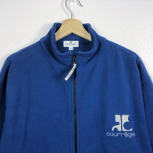 May include: A blue fleece jacket with a high collar and a full-length zipper. The jacket features the brand name "courr&egrave;ges" in white lettering on the lower right side. The jacket is hanging on a wooden hanger.