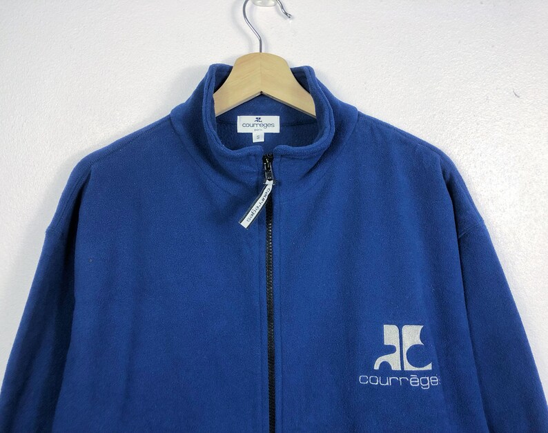 May include: A blue fleece jacket with a high collar and a full-length zipper. The jacket features the brand name "courr&egrave;ges" in white lettering on the lower right side. The jacket is hanging on a wooden hanger.