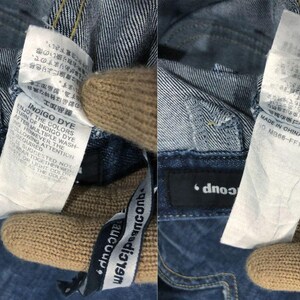 Op de afbeelding: Close-up van denimstof met aangehechte labels. E&eacute;n label luidt "Indigo Dye" met wasvoorschriften. Het andere label zegt "Made in China". De merknaam "dnooup" is zichtbaar.