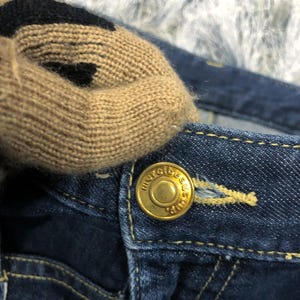 Op de afbeelding: Close-up van een donkerblauwe spijkerbroek met een gouden knoop. Een beige en zwart gebreid item ligt op de jeans. De knop is voorzien van de tekst "merci beau coup".
