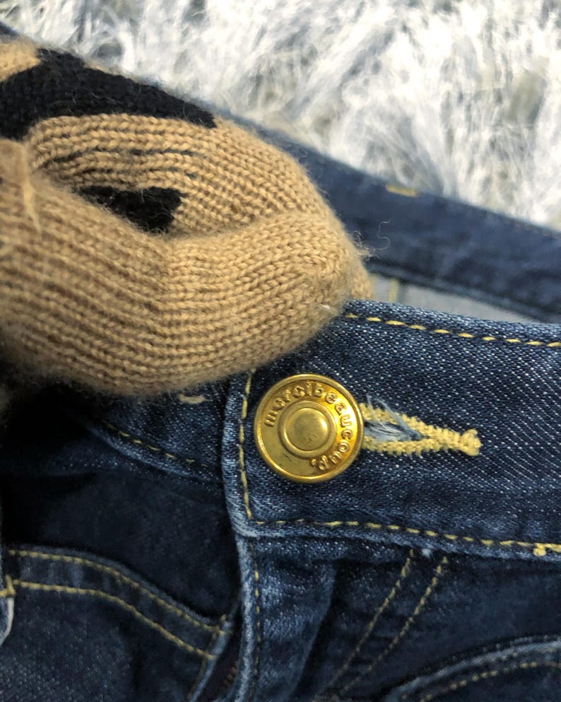 Op de afbeelding: Close-up van een donkerblauwe spijkerbroek met een gouden knoop. Een beige en zwart gebreid item ligt op de jeans. De knop is voorzien van de tekst "merci beau coup".