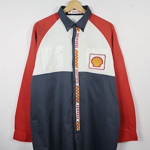 May include: A long-sleeve button-down shirt featuring a color-blocked design in red, white, and dark gray. The front displays a Shell logo patch on the chest and a vertical strip along the button placket with the text Shell and a checkered pattern.