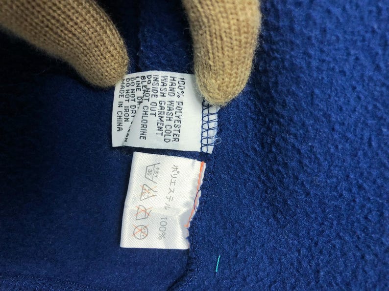 May include: Close-up of a blue fabric with attached care labels. The labels provide washing instructions, including hand wash cold, do not bleach, and do not iron. The fabric is made of 100% polyester and made in China.