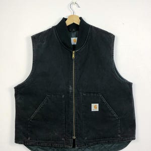 May include: A black Carhartt work vest with a zip fastening and a Carhartt logo patch on the front. The vest has two large pockets on the front and a quilted lining.