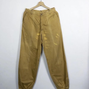 Op de afbeelding: Mosterdkleurige broek met een knoopsluiting en een losse pasvorm. De broek heeft een licht vervaagde uitstraling met enkele gele vlekken. De manchetten zijn bij de enkels gerimpeld. De broek hangt aan een houten kleerhanger.