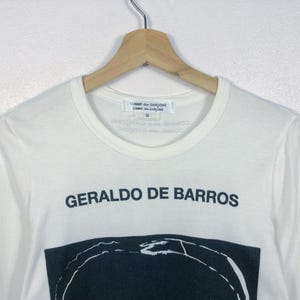 K&ouml;nnte beinhalten: Wei&szlig;es Langarmshirt mit dem schwarzen Text "GERALDO DE BARROS". Darunter befindet sich ein schwarz-wei&szlig;es Grafikdesign. Das Shirt h&auml;ngt an einem Holzkleiderb&uuml;gel.
