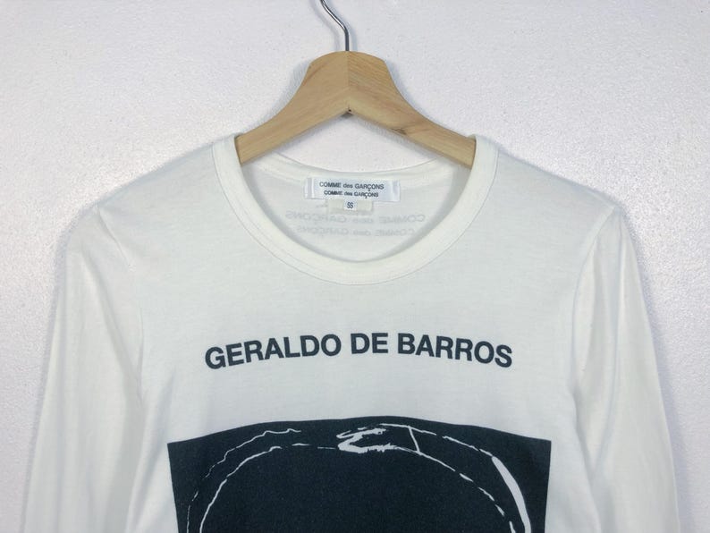 K&ouml;nnte beinhalten: Wei&szlig;es Langarmshirt mit dem schwarzen Text "GERALDO DE BARROS". Darunter befindet sich ein schwarz-wei&szlig;es Grafikdesign. Das Shirt h&auml;ngt an einem Holzkleiderb&uuml;gel.