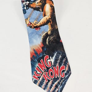 May include: A necktie featuring a vintage King Kong design. The tie depicts the giant ape holding a woman and battling airplanes, with the words "KING KONG" in red. The background is a blue sky with a cityscape.