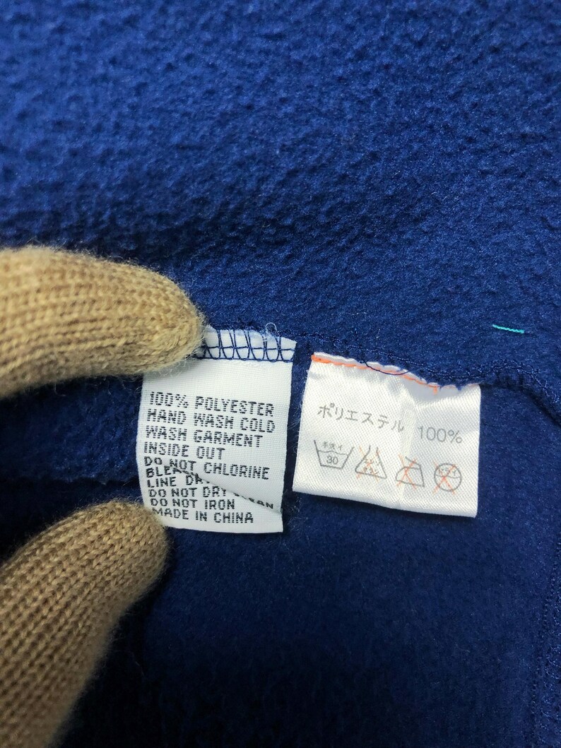May include: Close-up of a blue fleece fabric with two white tags. One tag reads "100% POLYESTER" and care instructions. The other tag has Japanese text and washing symbols. The fabric is made in China.