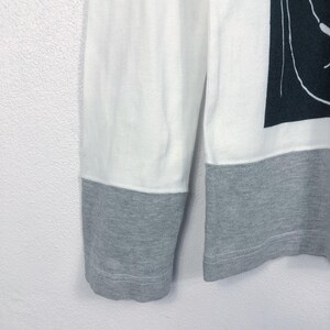 K&ouml;nnte beinhalten: Wei&szlig;es und graues Langarmshirt. Der obere Teil des Shirts ist wei&szlig;, w&auml;hrend die unteren &Auml;rmel und Manschetten grau sind. Auf der rechten Seite des Shirts ist eine schwarz-wei&szlig;e Grafik zu sehen.