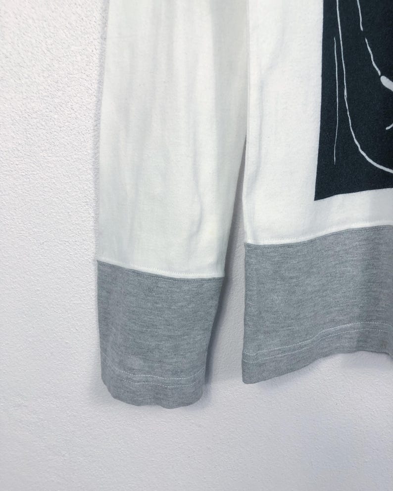 K&ouml;nnte beinhalten: Wei&szlig;es und graues Langarmshirt. Der obere Teil des Shirts ist wei&szlig;, w&auml;hrend die unteren &Auml;rmel und Manschetten grau sind. Auf der rechten Seite des Shirts ist eine schwarz-wei&szlig;e Grafik zu sehen.