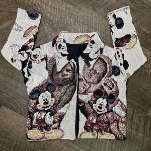 May include: A cream-colored jacket featuring a repeating pattern of Mickey Mouse and other Disney characters. The jacket has a collared neckline, black lining, and long sleeves. The design incorporates shades of brown, red, and blue.
