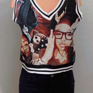 Pode incluir: Um colete sem mangas com decote em V com um design de tapeçaria colorido com retratos. O colete tem acabamento listrado em branco e preto. A modelo também usa shorts pretos. Peça de roupa moderna.