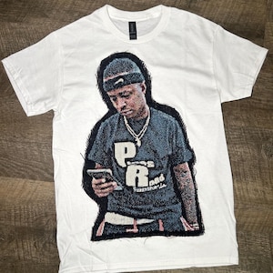 May include: White t-shirt with a detailed graphic of a person wearing a headband and holding a phone. The graphic includes the letters "P" and "R" with additional text. The shirt has a crew neck and short sleeves.