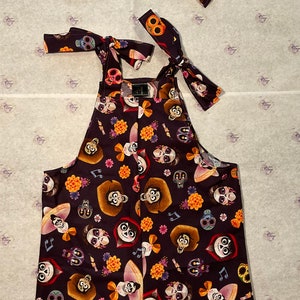 May include: A dark purple overall with a pattern of colourful flowers, skulls, and characters from the film Coco. The overall has two matching bow ties with the same pattern.