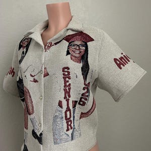 Pode incluir: Camisa bege com botões com um design tecido com tema de formatura. A camisa tem mangas curtas e gola. O design inclui imagens de uma graduada usando um chapéu e o texto "SENIOR 2023" em vermelho.
