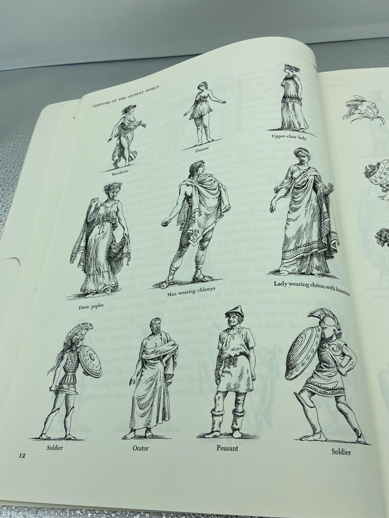 May include: An open book page featuring black and white illustrations of ancient world costumes. The illustrations include figures labeled as a dancer, upper-class lady, man wearing chlamys, lady wearing chiton, soldier, orator, and peasant.