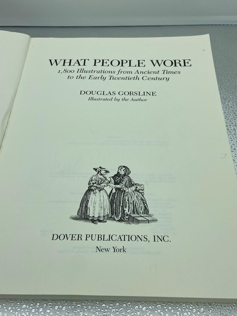 May include: Open book page with the title "WHAT PEOPLE WORE" and the subtitle "1,800 Illustrations from Ancient Times to the Early Twentieth Century." The author is Douglas Gorsline. A black and white illustration of two women in historical clothing is at the bottom.