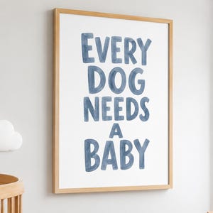 Op de afbeelding: Een rechthoekige wandkunstprint in een lichthouten lijst met de blauwe aquarel-stijl tekst EVERY DOG NEEDS A BABY gecentreerd op een witte achtergrond. De letters zijn in een vet, afgerond schreefloos lettertype met kleurvariaties.