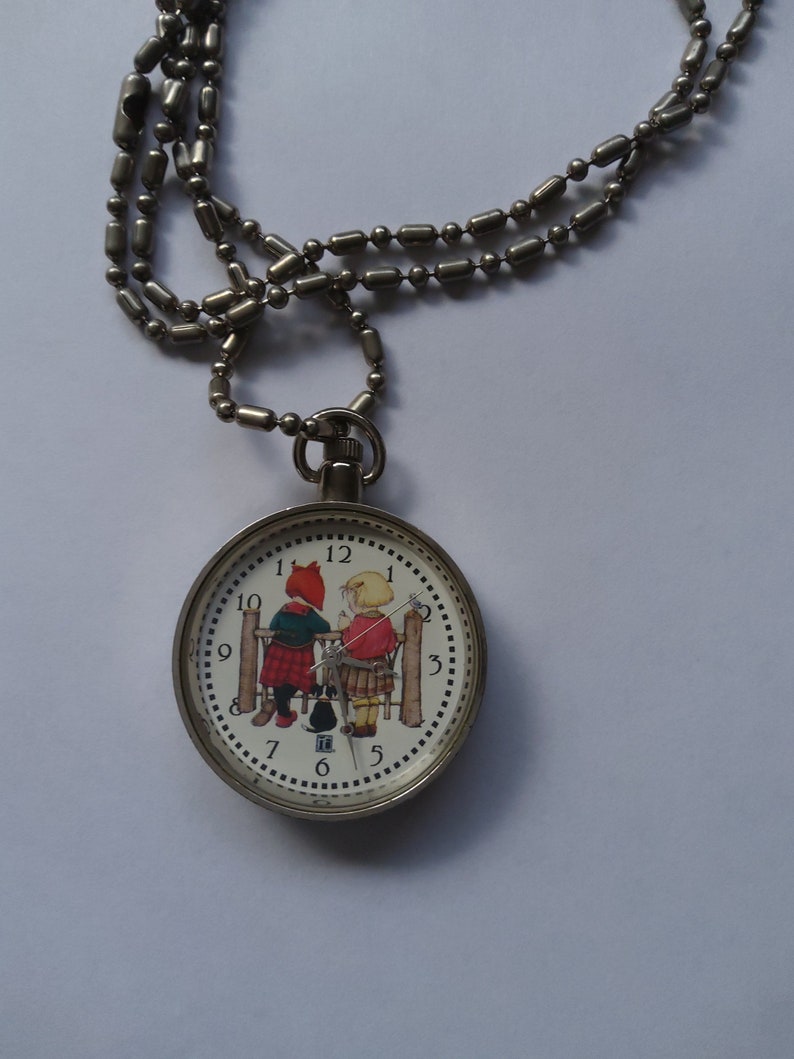 May include: A silver chain necklace with a round pocket watch pendant. The watch face features a white background with black numbers and a colorful illustration of two children standing by a fence.