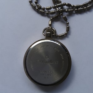 May include: Silver pocket watch with a chain. The back of the watch has the text "Mary Engelbreit" and "Concept in Time NY".