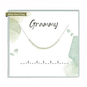 May include: A silver necklace with gold accents is displayed on a white card with the word "Grammy" in cursive. The card has watercolor-style green and gray accents. The necklace appears to spell out a message in Morse code.