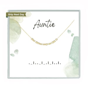 May include: A gold necklace with a delicate chain and small gold and silver beads. The necklace is displayed in a white box with the word "Auntie" printed in black and the text "..._..._..._..._..." printed below.
