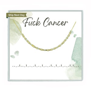 May include: A gold-colored necklace with a Morse code design is displayed on a white card. The card has the words "Fuck Cancer" in black script. The necklace is composed of small beads and cylindrical bars. The card has a watercolor design.