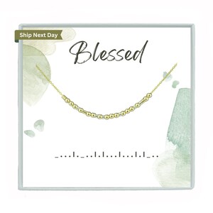 May include: A gold necklace with the word "Blessed" written on a white card with a green watercolour background.