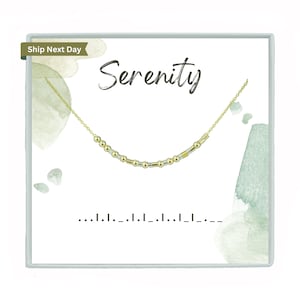 May include: A gold necklace with a delicate chain and small gold and silver beads. The necklace is displayed in a white box with the word "Serenity" written in black cursive lettering.