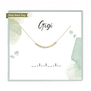May include: A gold-colored necklace with a delicate chain and a series of beads and bars. The necklace is displayed on a white card with the name "Gigi" in cursive and a Morse code pattern. Watercolor-style green and blue accents frame the design.