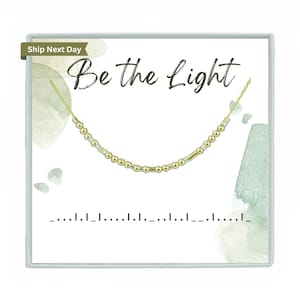 May include: A gold necklace with small beads and a bar charm. The necklace is in a white box with the words "Be the Light" written in black.