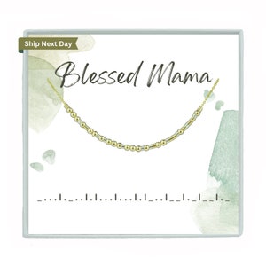 May include: A gold-coloured necklace with a beaded design, presented on a white card with the words "Blessed Mama" in elegant script. The card has watercolour-style green accents and a line of dashes and dots.