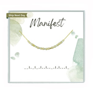 May include: A gold-colored necklace with a Morse code design, presented on a white card with the word "Manifest" in cursive script. The card has watercolor-style green accents and a light blue border.