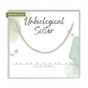 May include: A white card with a light green watercolor background. The card says "Unbiological Sister" in black cursive text. A silver and gold beaded necklace is displayed on the card.