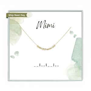 May include: A gold necklace with a delicate chain and small gold beads. The necklace is displayed in a white box with the word "Mimi" written in black cursive. The box has a green watercolor background.