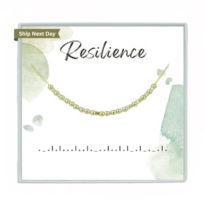 Peut inclure: Une boîte blanche avec un fond aquarelle vert et le mot "Résilience" en écriture noire. Un collier de perles dorées et argentées est présenté dans la boîte.