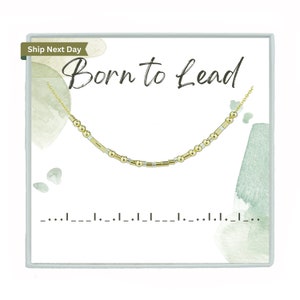 May include: A gold necklace with a Morse code pattern is presented on a white card. The card features the words "Born to Lead" in black script. The necklace is made up of small beads and rectangular bars. The background has watercolour-style green and grey accents.