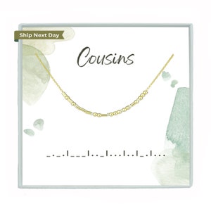 May include: A gold necklace with the word "Cousins" on a white card with a watercolor background. The necklace is made of small gold beads and has a delicate chain.