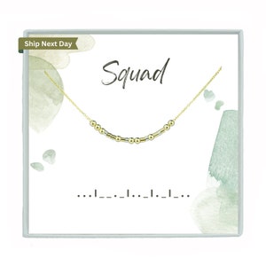 May include: A gold necklace with a Morse code message, displayed on a white card. The word "Squad" is written in a script font above the necklace. The background features watercolor-style green and grey accents. The necklace is a delicate piece of jewellery.