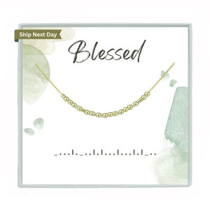 May include: A gold-coloured beaded necklace with a delicate chain, displayed on a white card. The card features the word "Blessed" in elegant script and watercolour accents. The necklace is designed with a Morse code pattern.