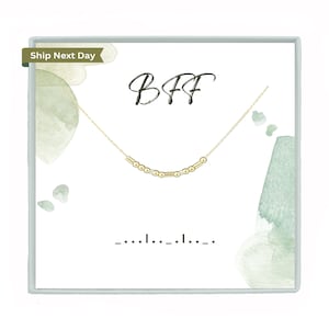 May include: A gold-colored necklace with a Morse code design, presented on a white card with the word "BFF" in cursive script. The card has watercolor-style green accents. The Morse code spells out "BFF".